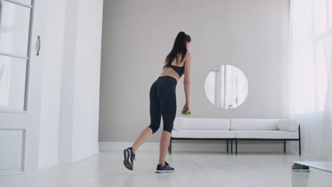 Young-beautiful-brunette-woman-standing-on-one-leg-doing-bends-with-dumbbells.-Performing-deadlift-with-dumbbells-at-home.